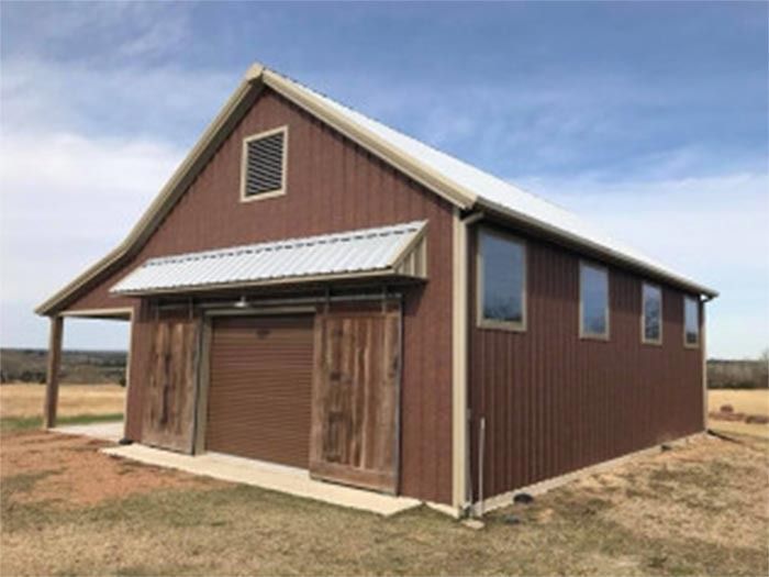 A brown barn with a metal roof and sliding wooden doors set in a dry, grassy field under a blue sky.