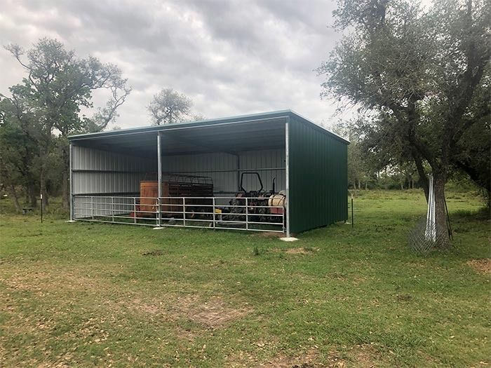 A dark green metal pole barn stands in a grassy field, housing farm equipment and a fenced area for livestock.
