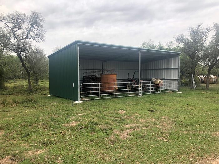 A green and white metal open-sided shed stands in a grassy field with equipment stored inside behind metal fencing.