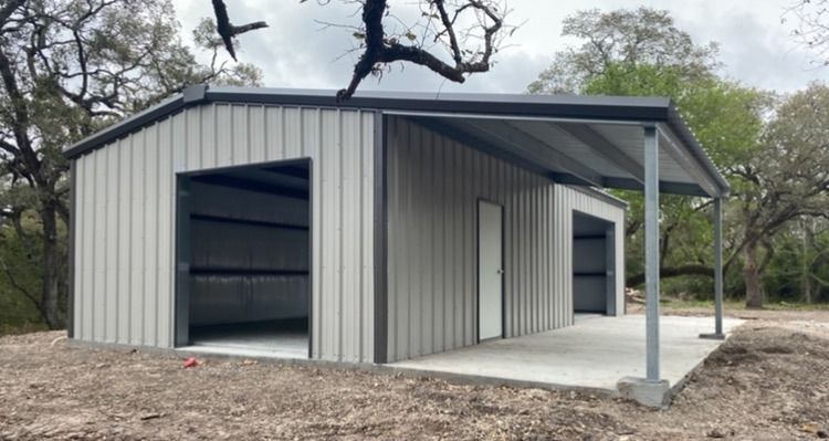 A gray metal building with an open garage bay and a side porch on a concrete slab in a wooded area.