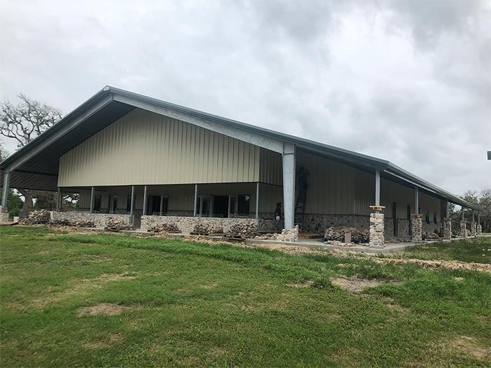 A modern, metal-framed building with a stone base and tan siding sits on a grassy lot under a cloudy sky.