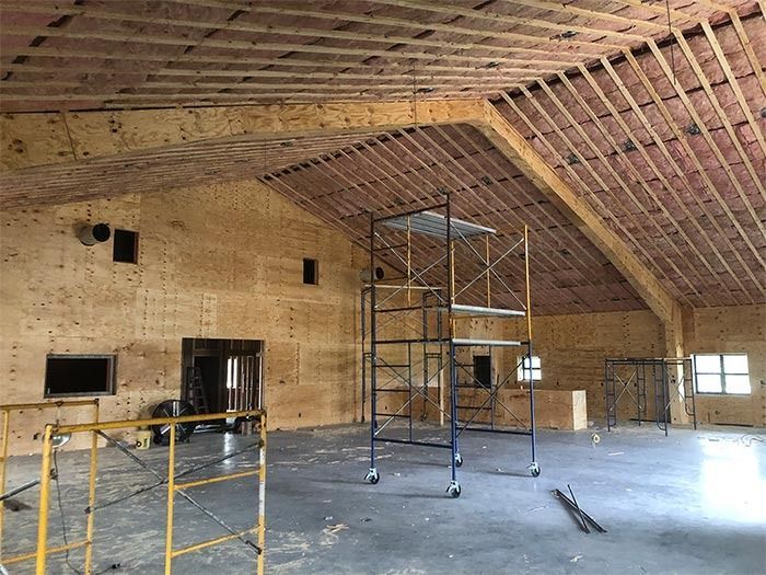 A wide, unfinished room under construction with plywood walls, exposed ceiling rafters, and metal scaffolding.