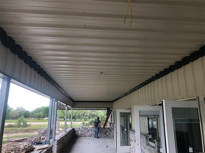 A person stands on a covered porch with a metal ceiling, facing an outdoor landscape under construction.