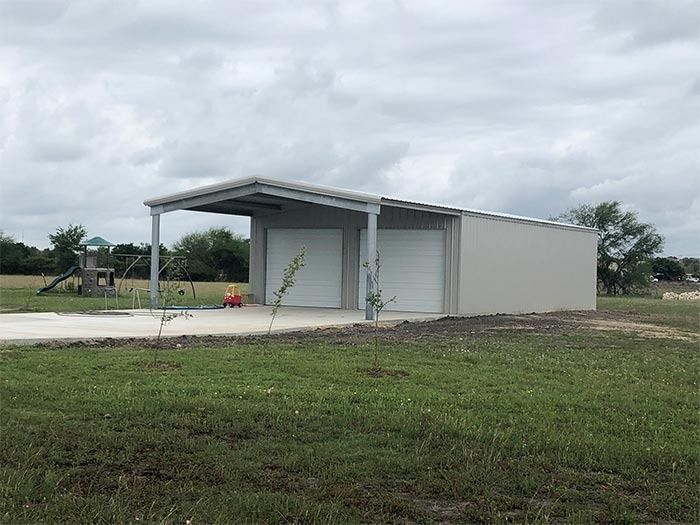 A gray metal garage with two roll-up doors and an open-sided carport area set on a grassy rural property.