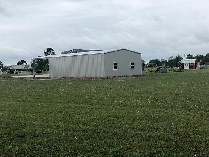 A gray metal barn with an open side porch stands in a large, grassy field under a cloudy sky.