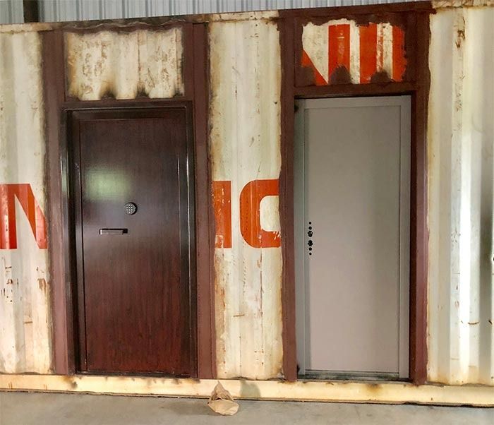Two doors installed in a weathered white and orange shipping container wall; one dark brown and one light grey.