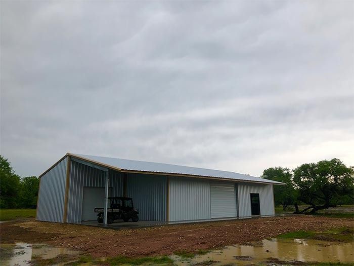A metal barn with a covered porch and an all-terrain vehicle parked inside, set on a gravel lot under a cloudy sky.