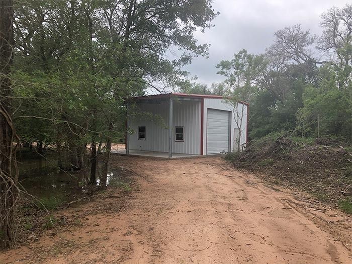 A white metal garage building with a red roof sits in a wooded area at the end of a dirt driveway.