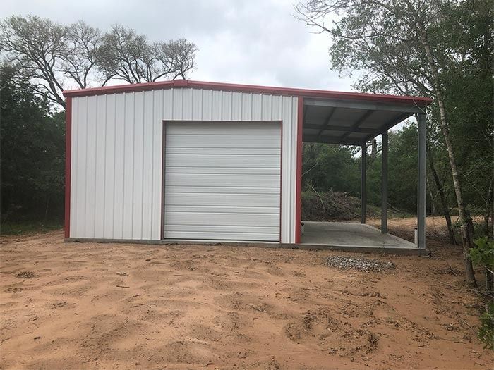 A white metal building with a garage door and an attached open-sided carport, situated on a sandy lot surrounded by trees.