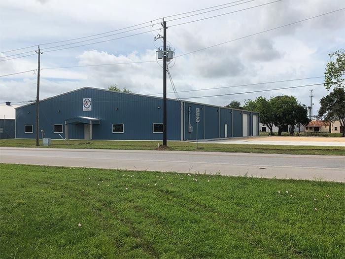 A blue, single-story industrial metal building sits behind a grassy field and a paved road under a cloudy sky.