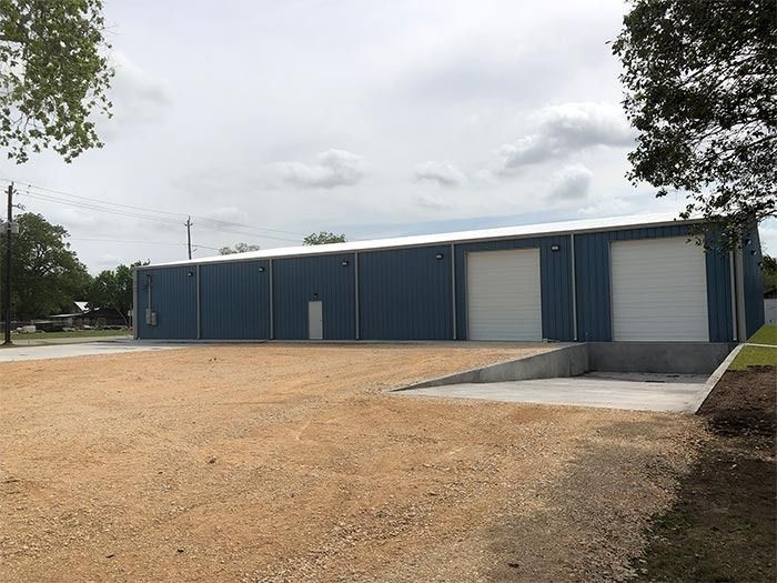 A blue, rectangular industrial warehouse with two white roll-up doors and a concrete loading ramp on a gravel lot.