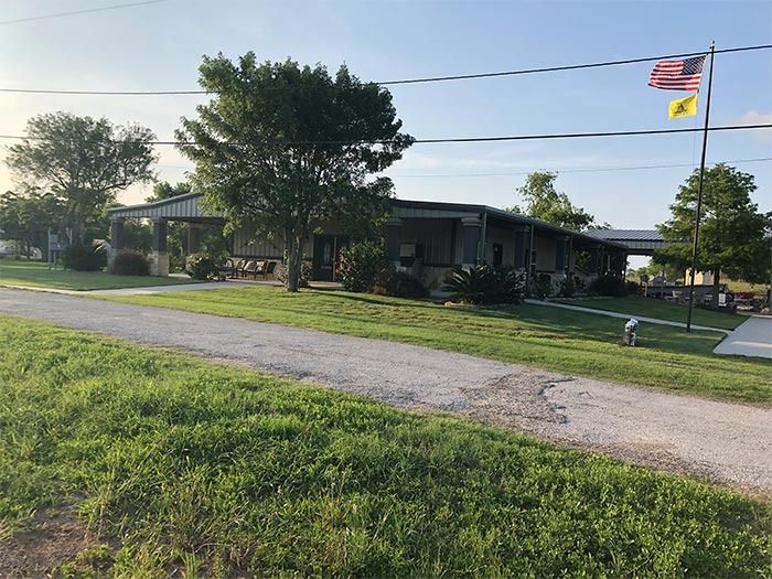 A single-story building with a covered entrance sits on a grassy lot with trees and a flag pole displaying two flags.