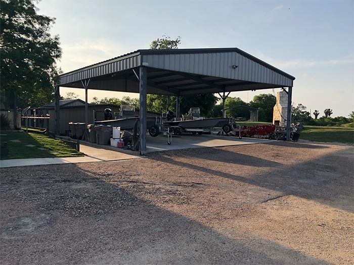 A large, gray, open-sided metal carport sits on a gravel lot during the daytime, housing a boat and trailer.