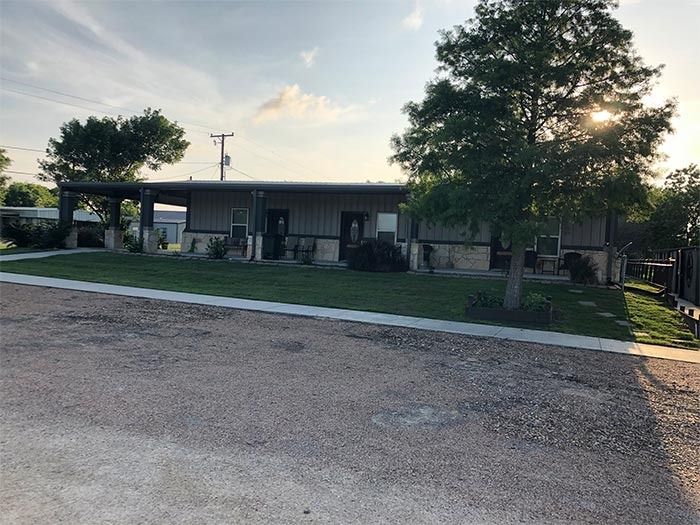 A single-story, neutral-colored commercial building with a covered porch and a grassy lawn, viewed from a gravel lot.