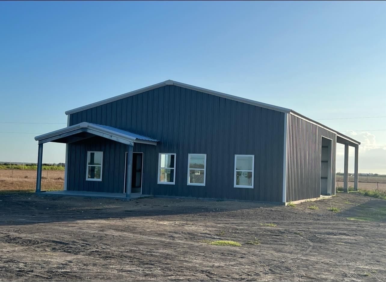 A dark grey metal building with a small covered porch entrance and several windows, situated in an open, dirt-filled field.