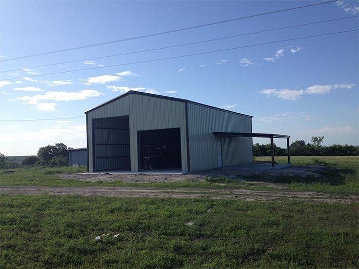 A light-colored metal workshop building with two open doorways and an attached side canopy in a grassy field.