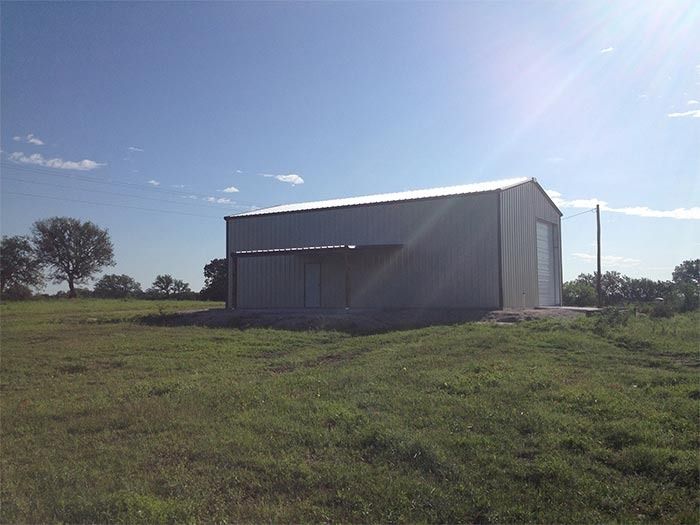 A metal pole barn with a silver roof sits in a grassy, open field under a sunny blue sky.