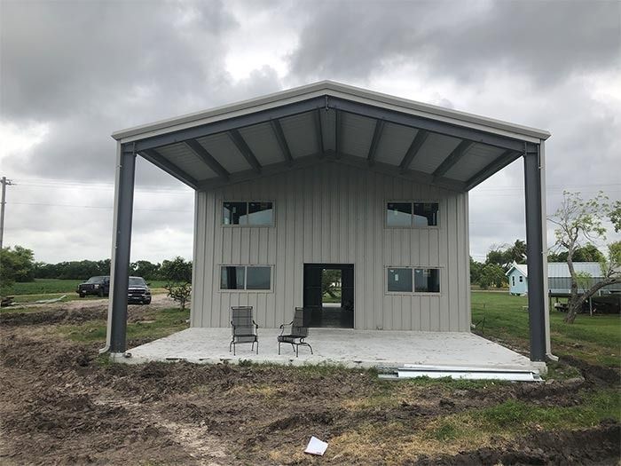 A two-story metal building with a large covered porch and gray siding set against a cloudy sky over a dirt yard.
