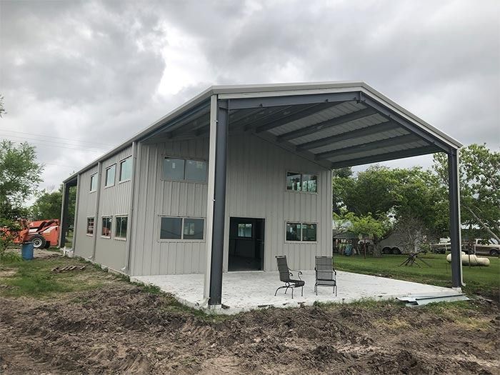 A two-story, grey metal building with an attached open-air carport featuring a concrete patio and two chairs.