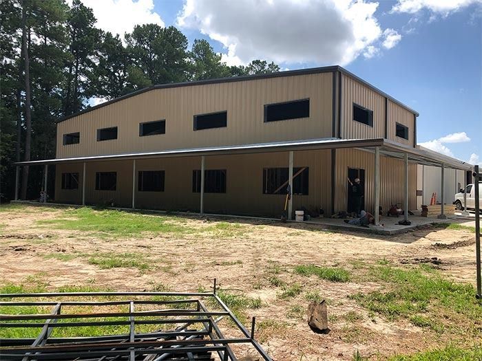 A beige two-story metal building under construction with a long, thin awning and dirt ground in a wooded area.