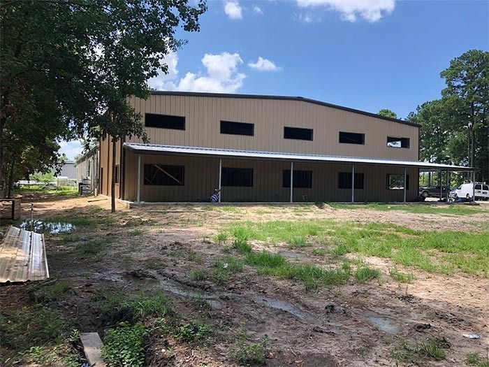 A tan metal building with dark windows and a covered porch, set on a muddy, grassy lot under a sunny blue sky.