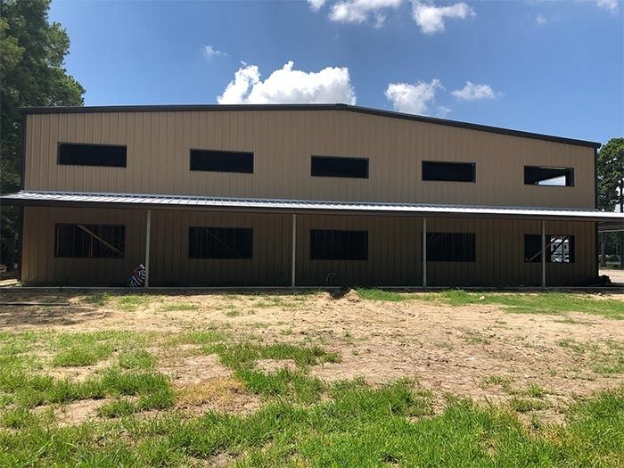A large, tan metal building under construction with two levels of empty window openings and a front porch, on grass.