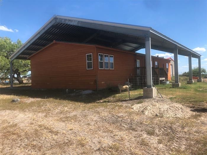 A brown mobile home sits underneath a large, open-sided metal carport structure on a sunny, dry grassy lot.