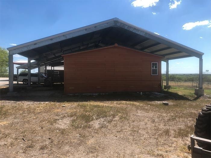 A brown, single-story house sheltered under a large, open-sided metal carport on a grassy, rural lot under a blue sky.