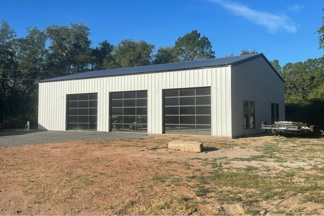 A white, modern metal workshop building with three glass-paneled garage doors, situated in a dirt and wooded lot.