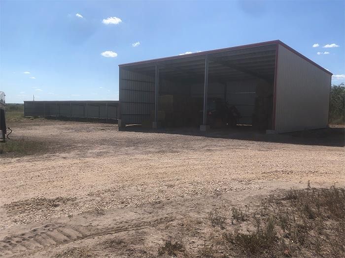 A light-colored, open-front metal barn sits on a gravel lot under a clear blue sky.