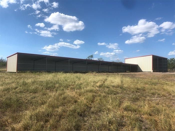 A long, beige metal shed with a sloping roof in a dry, grassy field under a bright, sunny blue sky.