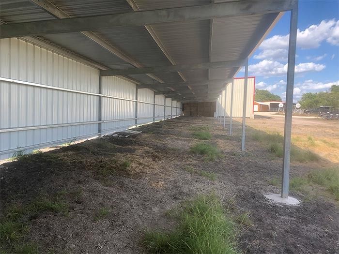 An outdoor metal-sided shed structure with a roof overhang and support posts, overlooking a dirt and grass area.