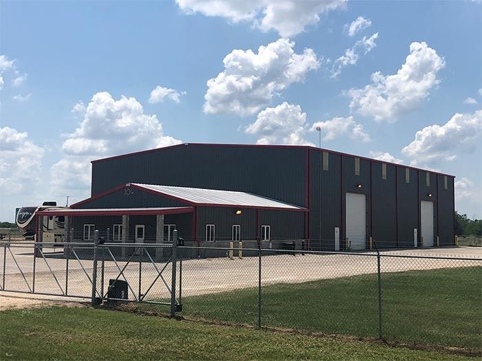 A dark gray industrial metal building with red trim, a white roofed porch, and two large garage doors, behind a fence.