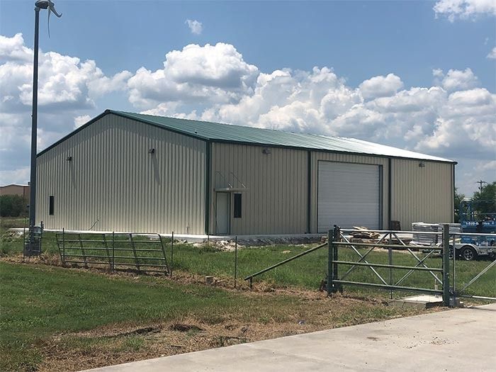 A tan metal warehouse with a green roof and a large garage door, situated on a grassy lot under a blue sky with clouds.