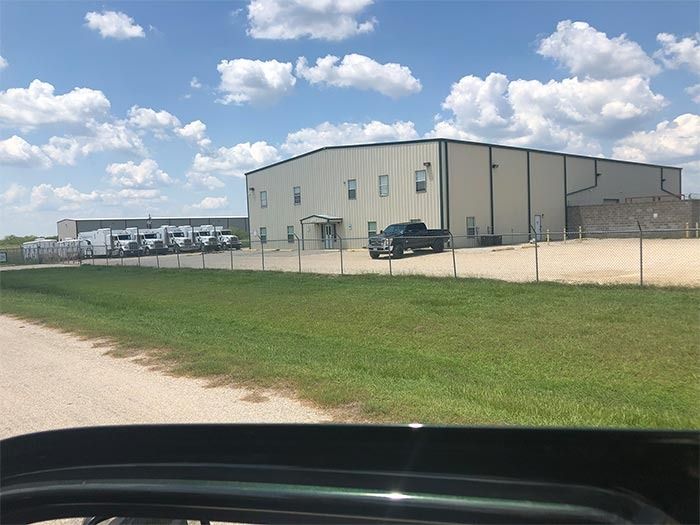 A beige commercial warehouse with a parked pickup truck, chain-link fence, and several trailers under a cloudy sky.