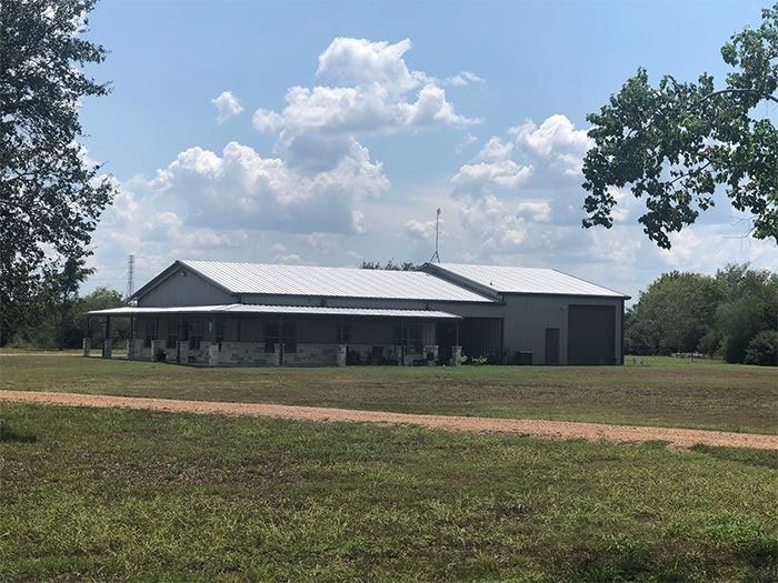 A gray, ranch-style home with a metal roof and large porch, situated on a grassy field under a sunny blue sky.