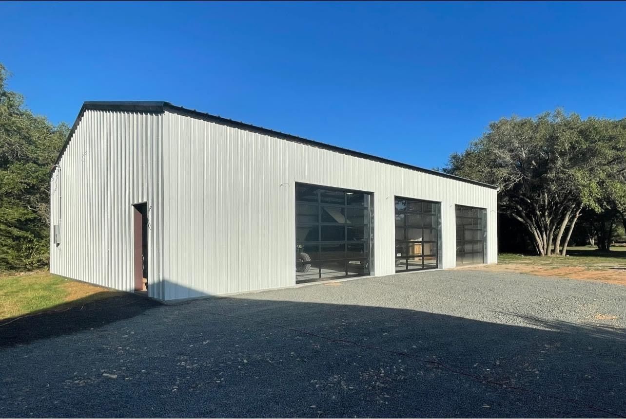 A white metal garage building with three glass-paneled garage doors and a side entry door, set on a gravel lot.
