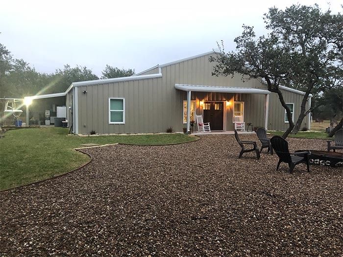 A tan metal-sided building with a covered entrance, rocking chairs, and patio furniture on a gravel yard at dusk.