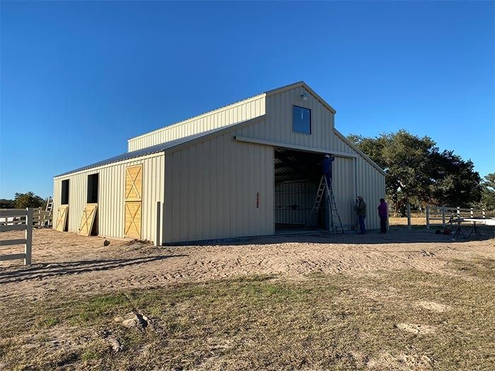 A beige metal barn with a central sliding door sits in a grassy, open lot under a clear blue sky.