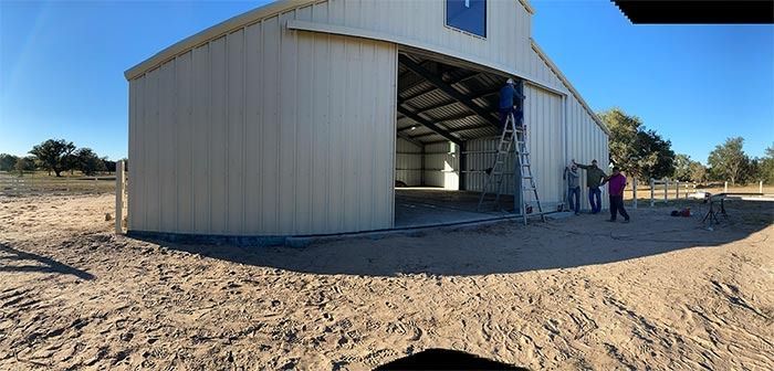 People install a large metal sliding door on a light-colored barn in an open, sandy rural area under a clear blue sky.