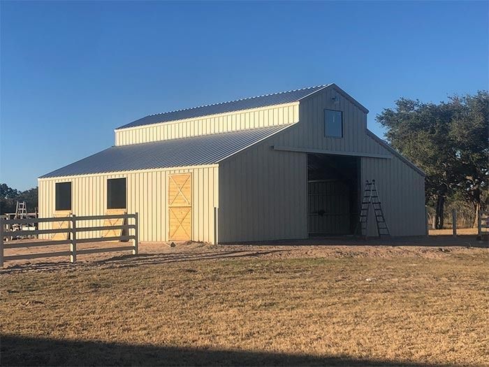 A beige metal barn with a raised center roof, a large open doorway, and a white fence in a grassy field under a blue sky.