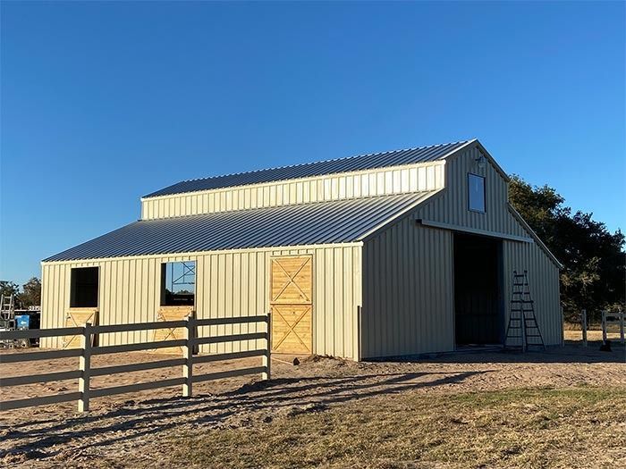 A beige, metal-sided barn with a dark roof and wooden sliding doors sits on a dirt lot beside a white fence.