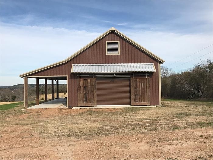 A brown metal barn with a front porch, sliding wooden doors, and a metal roof set in a grassy field under a blue sky.
