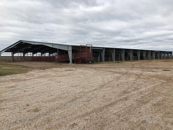 A long, open-sided metal livestock shelter sits on a dirt lot under a cloudy sky.