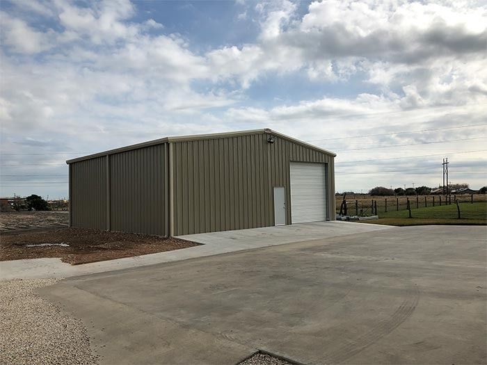 A beige metal storage building with a white roll-up door and a paved driveway under a cloudy sky.