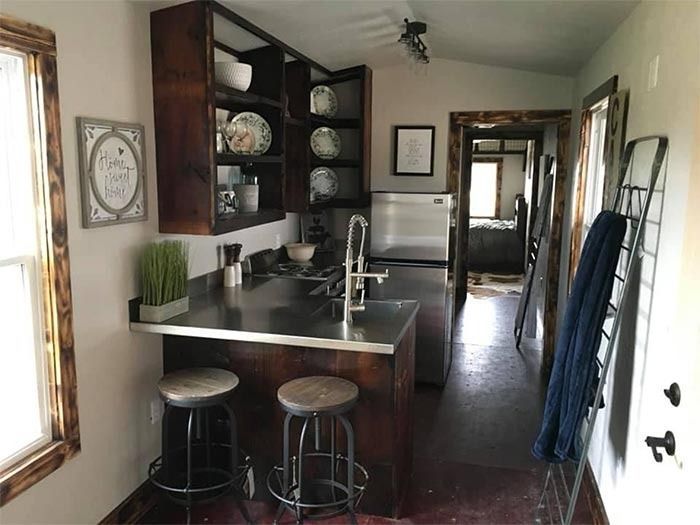 A rustic kitchen with wood cabinets, stainless steel counters, two bar stools, and an open doorway leading to a bedroom.