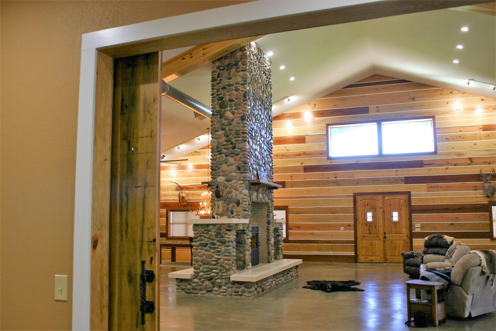 A rustic lodge-style room features a central stone fireplace, horizontal wood plank walls, and a large wooden door.