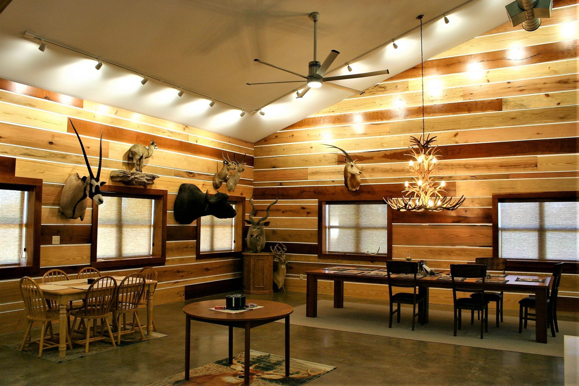 A rustic room with horizontal wood-paneled walls, taxidermy mounts, multiple dining tables, and an antler chandelier.