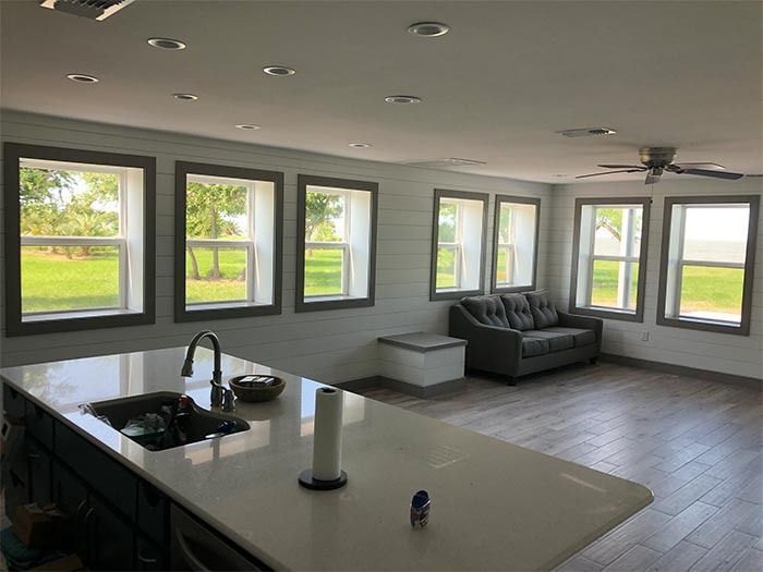 A kitchen island with a sink sits in the foreground of a room with light shiplap walls, a gray sofa, and several windows.
