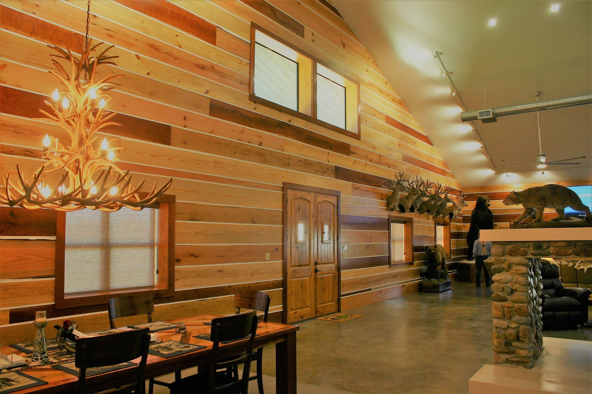 A rustic lodge interior with paneled walls, a large antler chandelier, a dining set, and a stone fireplace.
